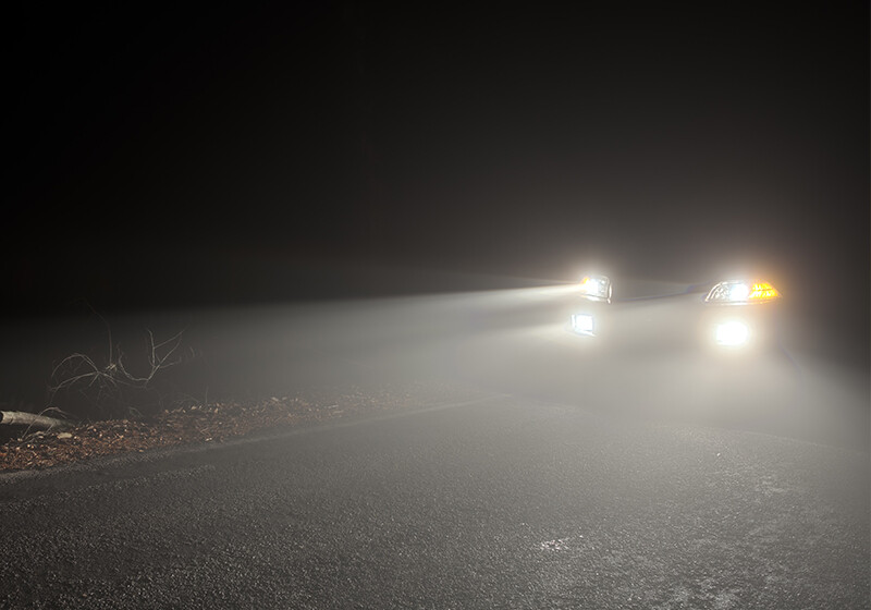 Ein Auto fährt nachts mit eingeschaltetem Abblendlicht durch dichten Nebel auf einer Landstraße.