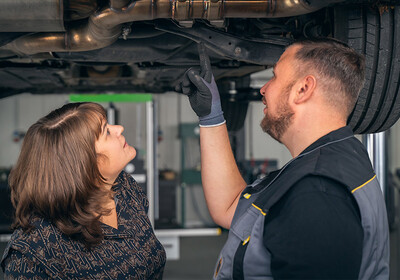 Eine Person in Arbeitskleidung zeigt einer anderen Person unter einem aufgebockten Auto einen bestimmten Bereich, offenbar im Rahmen einer technischen Beratung oder Fahrzeuginspektion. Im Hintergrund ist eine Werkstatt zu erkennen, die auf Autoreparaturen spezialisiert ist.