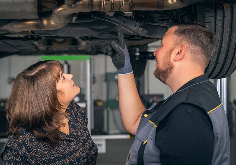 Eine Person in Arbeitskleidung zeigt einer anderen Person unter einem aufgebockten Auto einen bestimmten Bereich, offenbar im Rahmen einer technischen Beratung oder Fahrzeuginspektion. Im Hintergrund ist eine Werkstatt zu erkennen, die auf Autoreparaturen spezialisiert ist.
