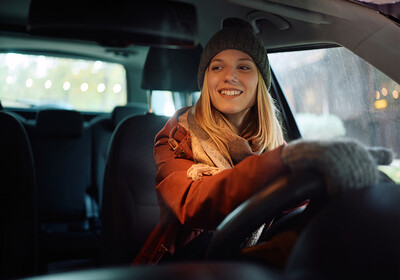 Eine lächelnde junge Frau mit blonden Haaren, grauer Strickmütze und rostbrauner Winterjacke sitzt am Steuer eines Autos. Die Aufnahme zeigt sie bei herbstlich-winterlichem Wetter, während sie entspannt nach vorne blickt.