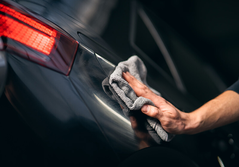 Eine Hand poliert mit einem Tuch die Karosserie eines dunklen Autos, das Rücklicht ist sichtbar.